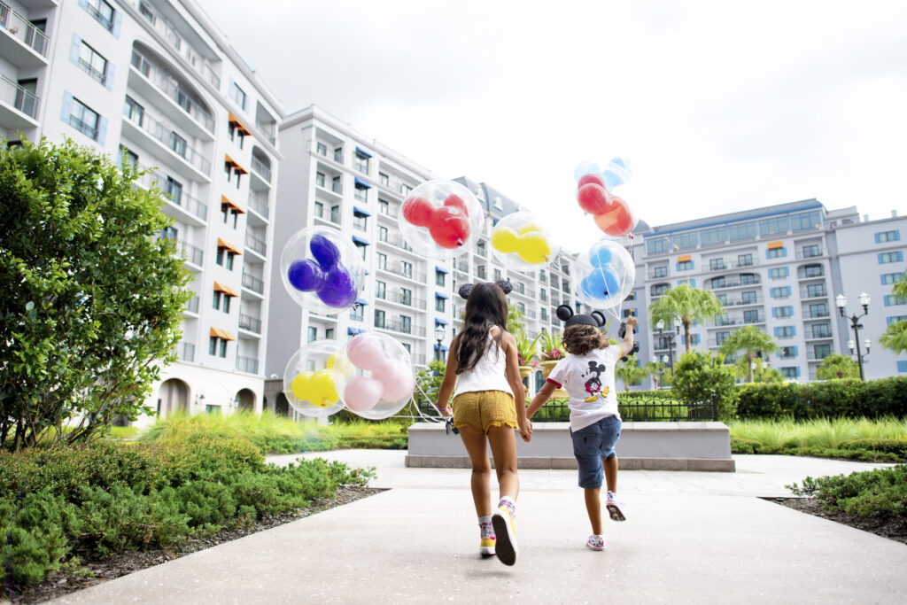 Two children run in the courtyard of Disney's Riviera Resort while holding colorful Mickey Balloons and wearing Minnie Mouse ears.