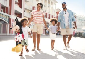 A happy and smiling family walks along Disney's Boardwalk while one child carries a large Mickey Mouse plush doll.