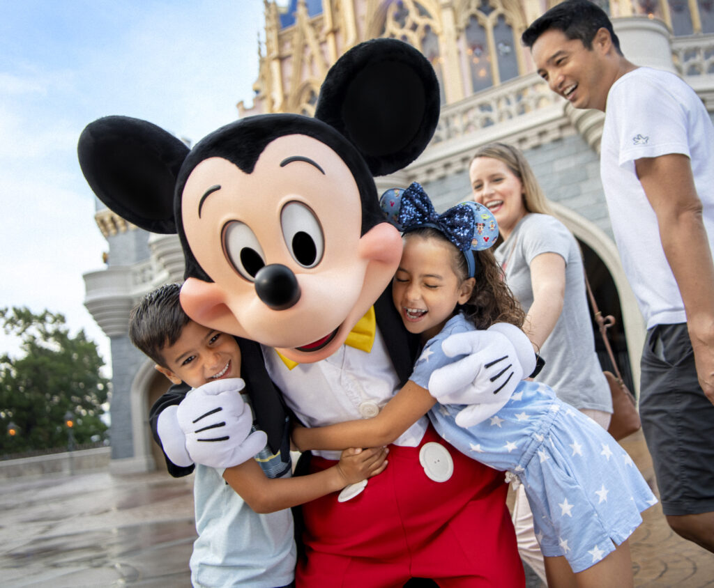 Two children smile and hug Mickey Mouse in front of Cinderella's Castle while their parents look on happily.
