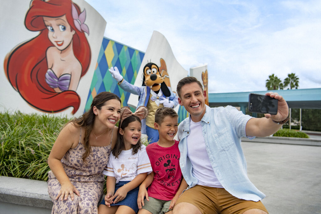 A happy and smiling family take a selfie outside of Disney's Art of Animation Resort at Walt Disney World while Goofy poses behind them.